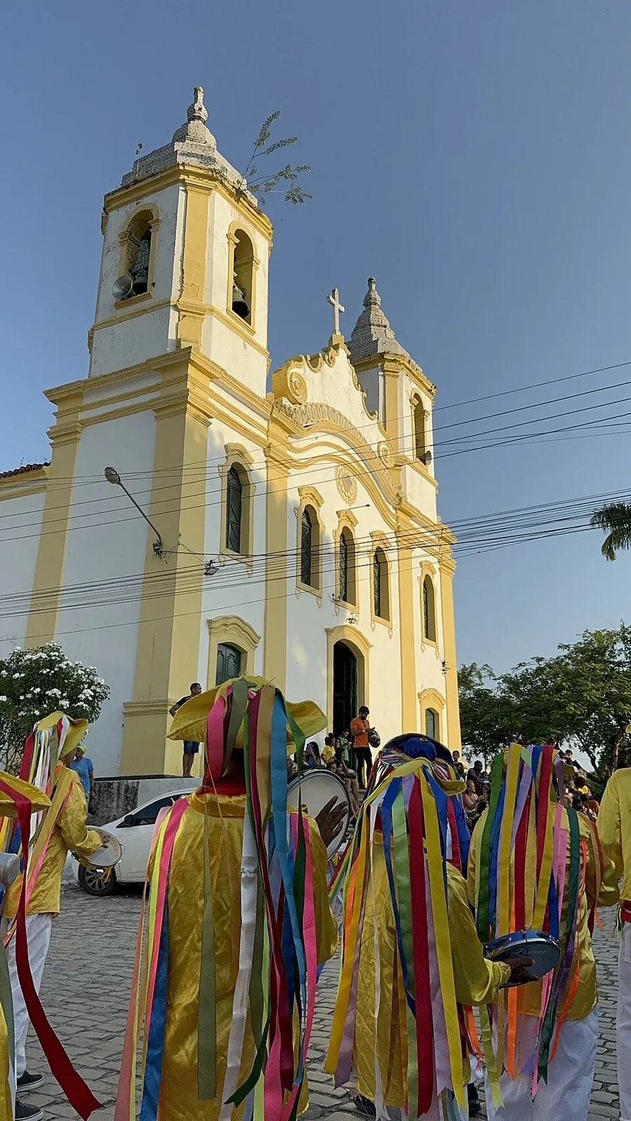 Integrantes do Cacumbi do Mestre Deca diante de igreja em Laranjeiras, com trajes amarelos e fitas coloridas